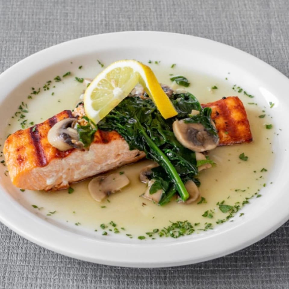 Grilled salmon fillet with sautéed spinach, mushrooms, and a lemon slice in a light sauce.