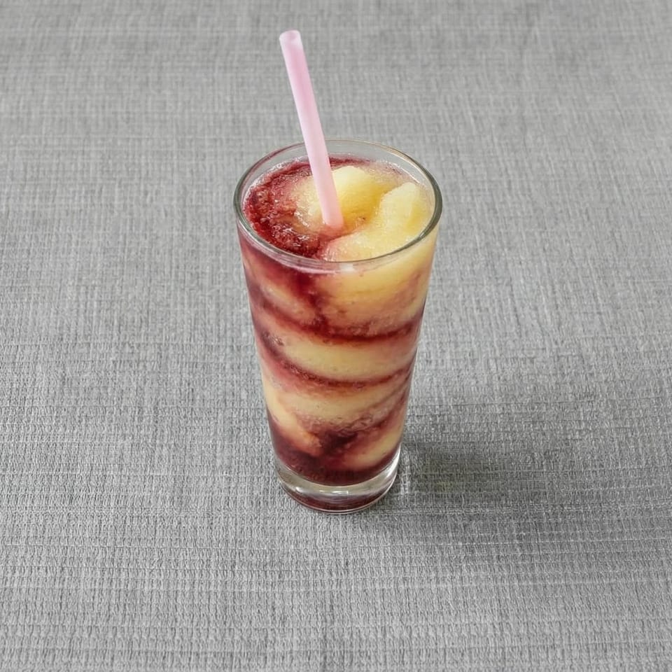 A tall glass of swirled red and yellow frozen drink with a pink straw on a gray surface.